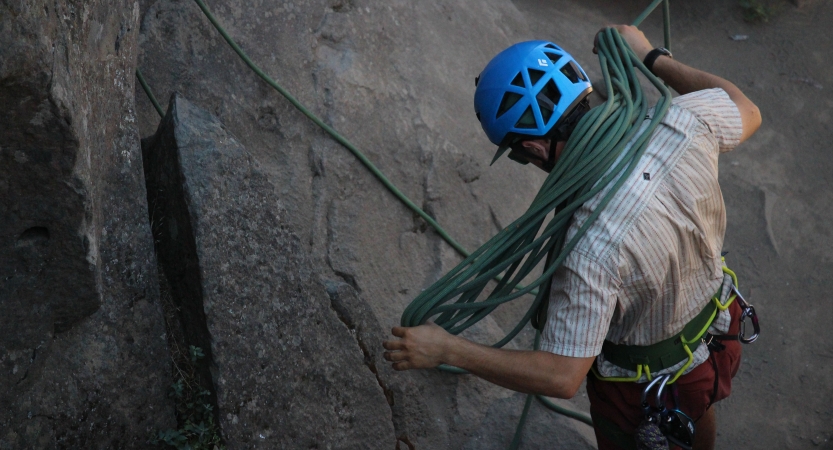 A person wearing safety gear uses their shoulders to fold a rock climbing rope while standing next to a rock wall